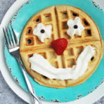 A waffle on a blue plate decorated with whipped cream and a strawberry to make a smiling face