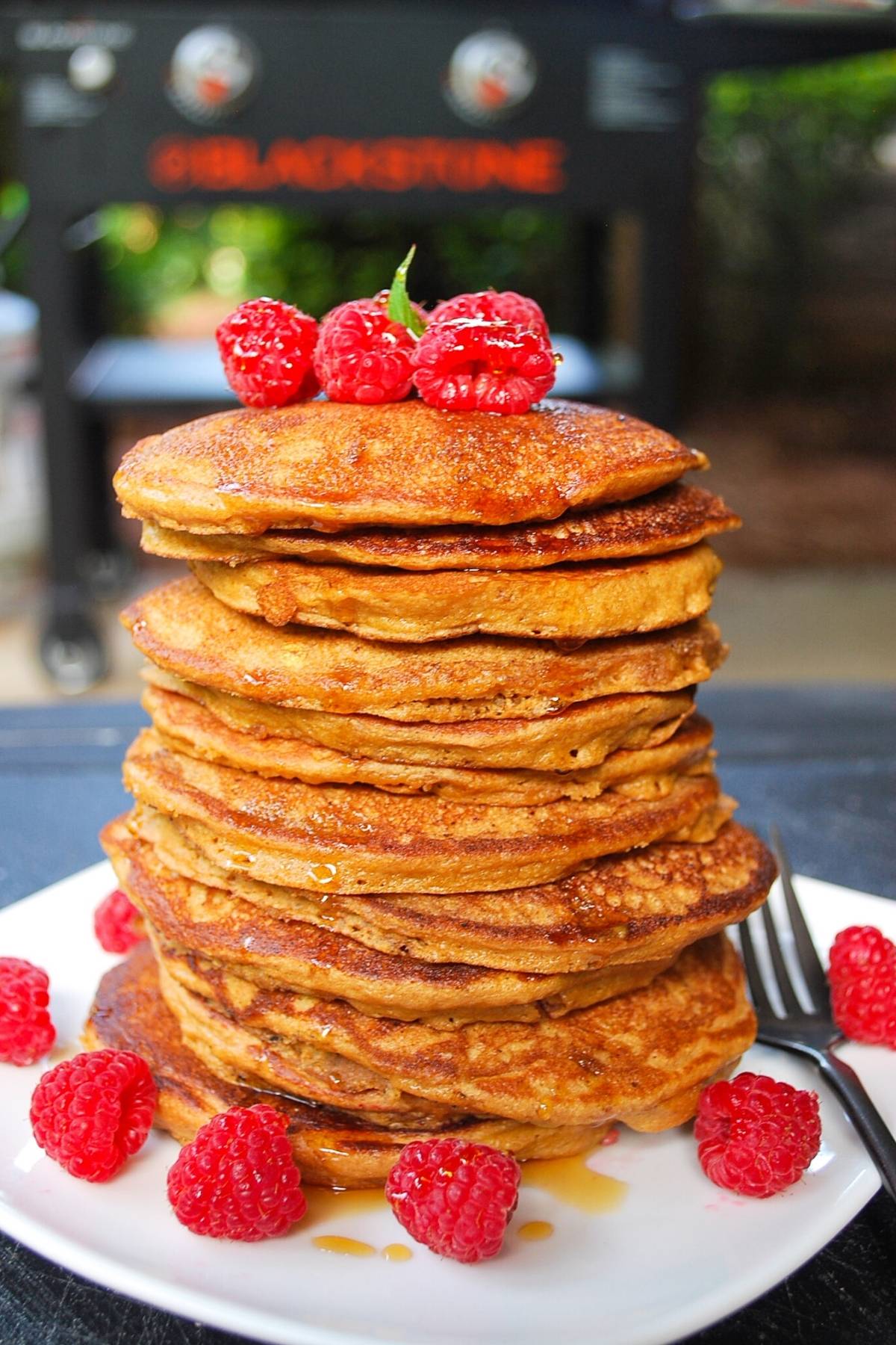 tall stack of buttermilk griddle pancakes topped with fresh raspberries and maple syrup