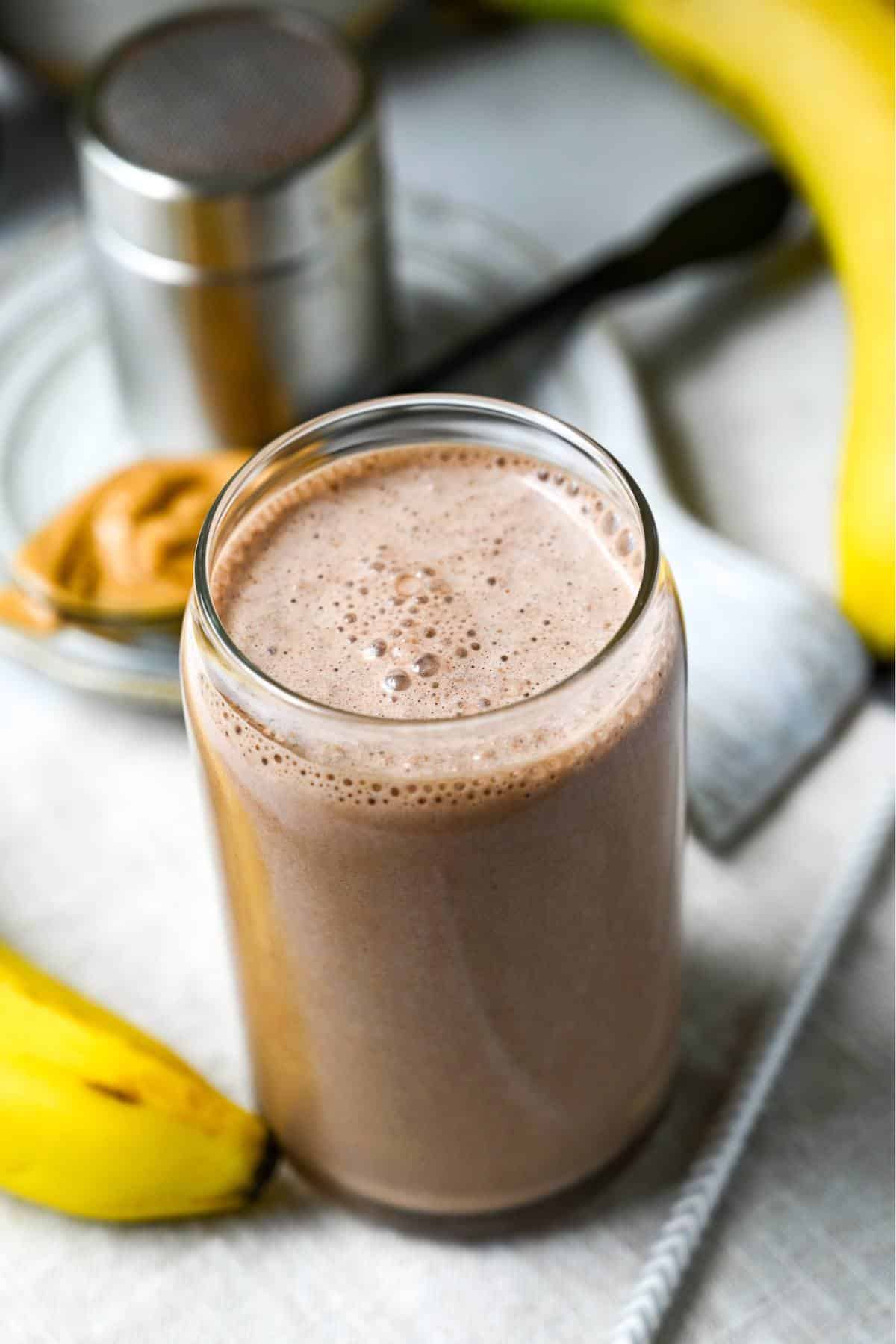 a chocolate protein shake in a glass with bananas and a spoonful of peanut butter next to it