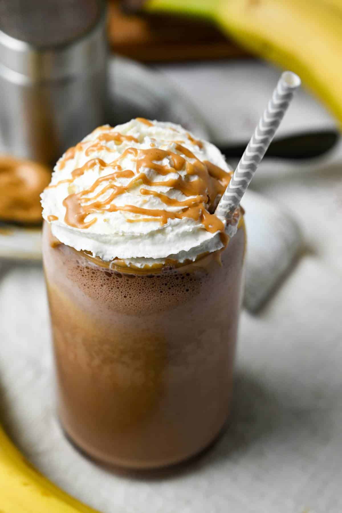 a chocolate pb banana protein shake in a glass topped with whipped cream and drizzled with melted peanut butter