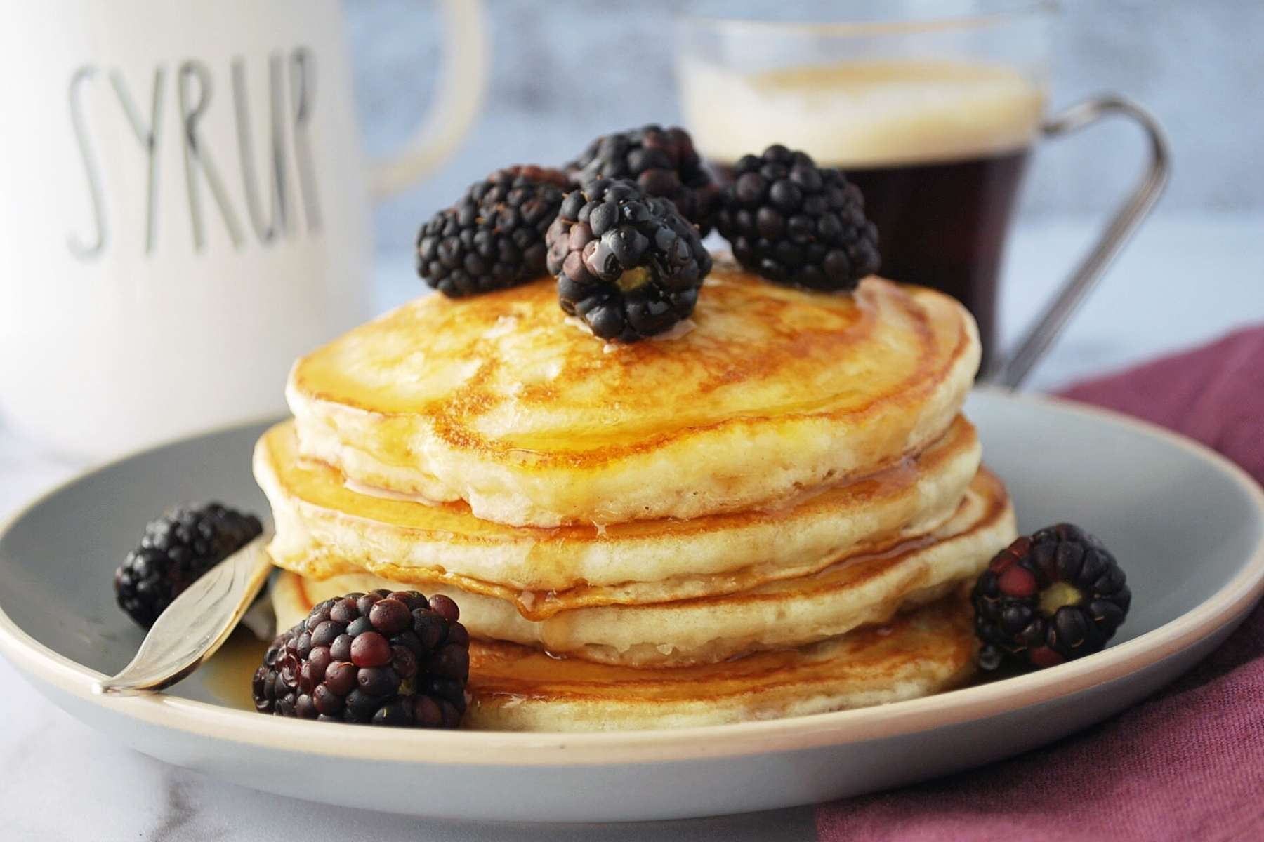 side view of a stack of pancakes with blackberries and syrup with a container of syrup and coffee in the background