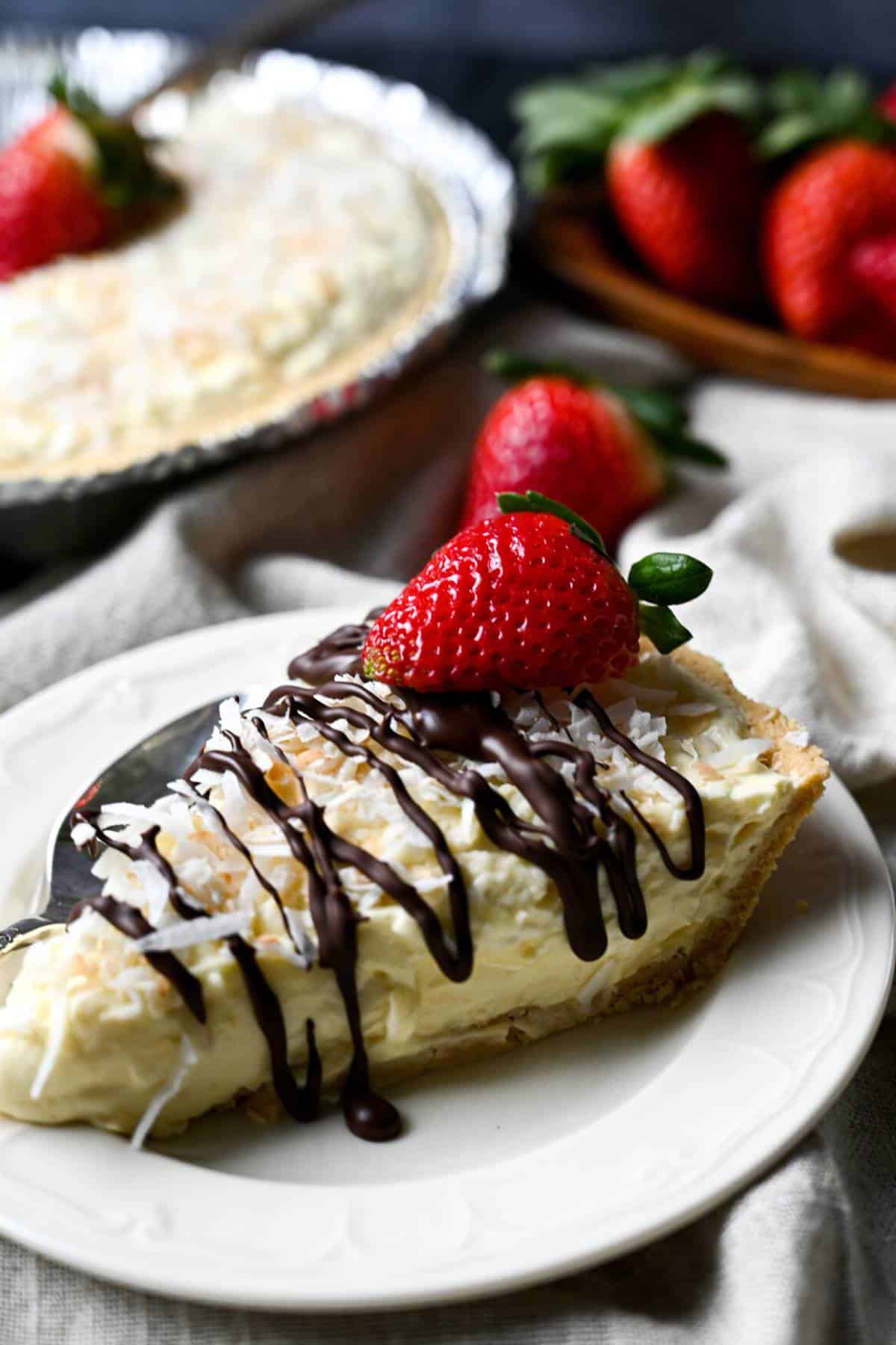 a slice of coconut cream pie drizzled with melted chocolate with a dish of fresh strawberries and whole pie behind it