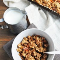 homemade granola in a cereal bowl and on a baking sheet and a small pitcher of milk