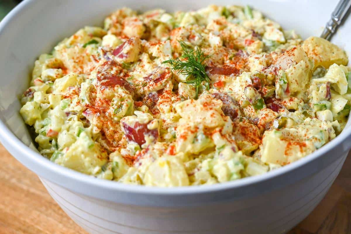 red potato salad in a white serving bowl with a spoon with a sprig of dill on top