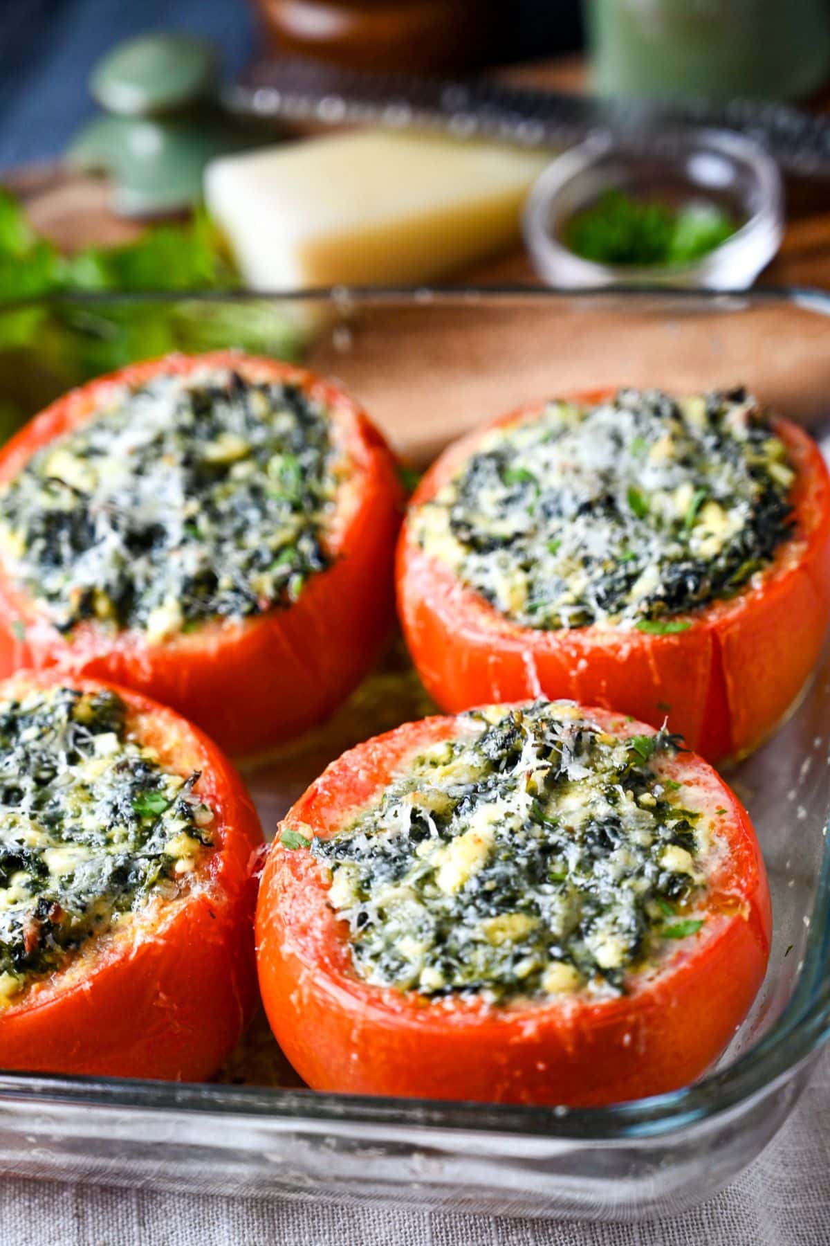 four spinach and cheese baked tomatoes with a cutting board behind them with a block of cheese, microplane, herbs, and pepper mill