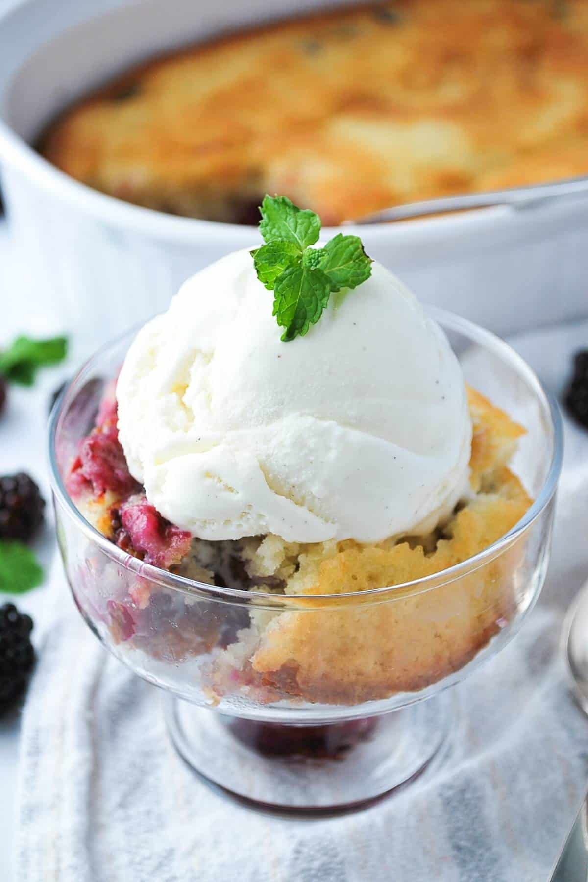 a dish of cobbler with ice cream in front of a baking dish of blackberry cobbler