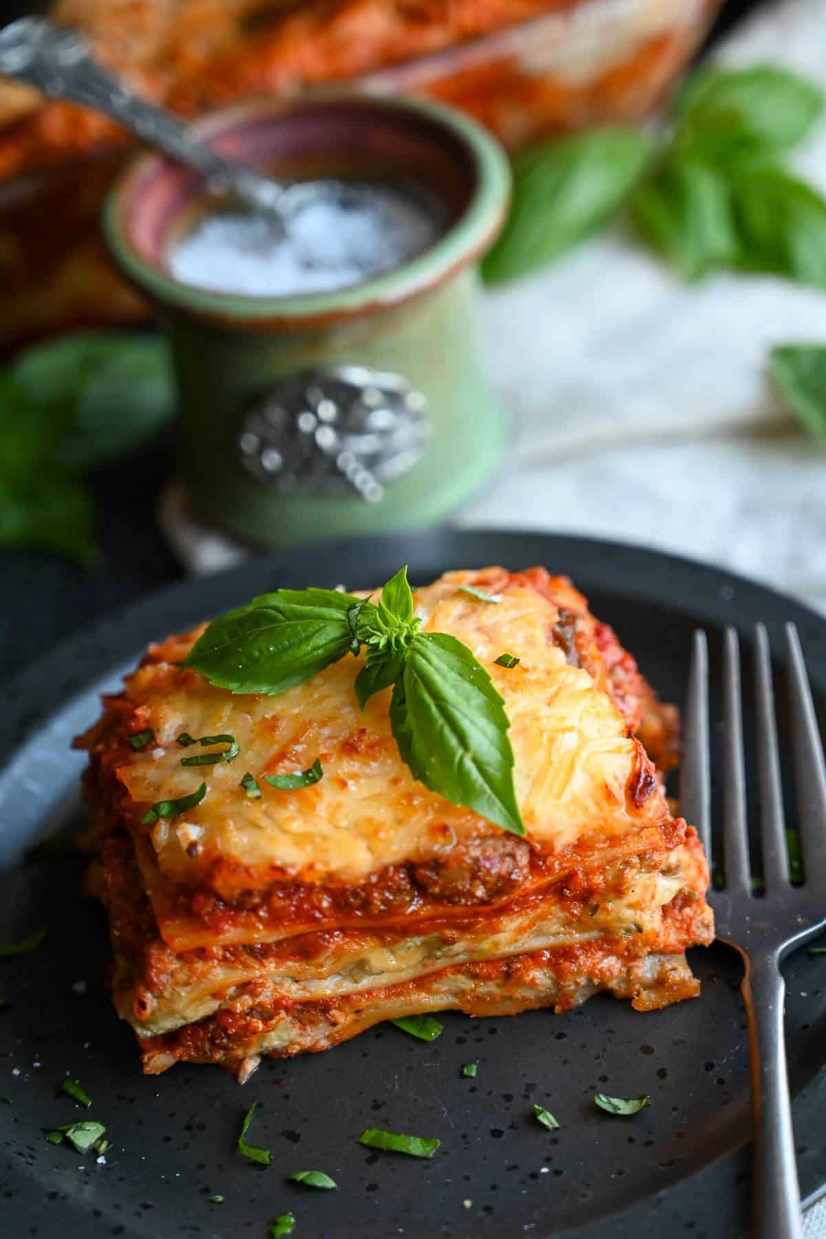a slice of lasagna on a black plate with a sprig of basil on top and a fork and salt container next to it