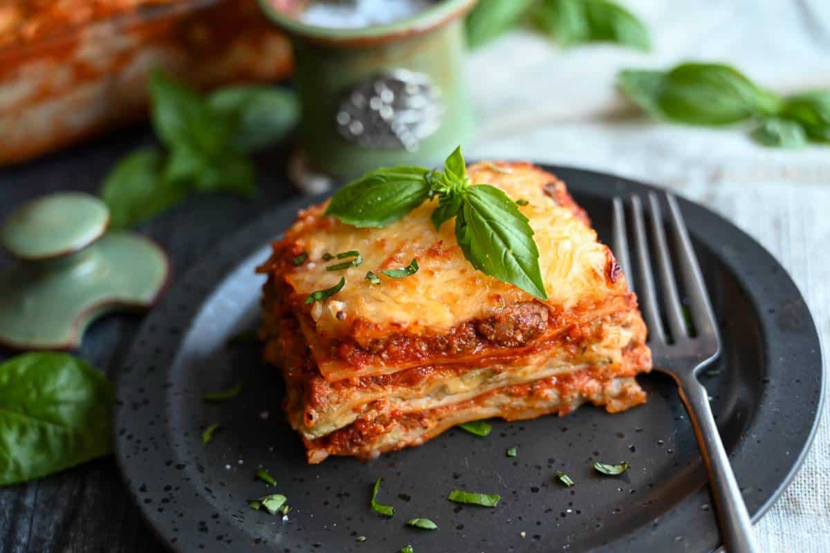 a sliced serving of lasagna with pesto sauce on a black plate with a fork and fresh basil