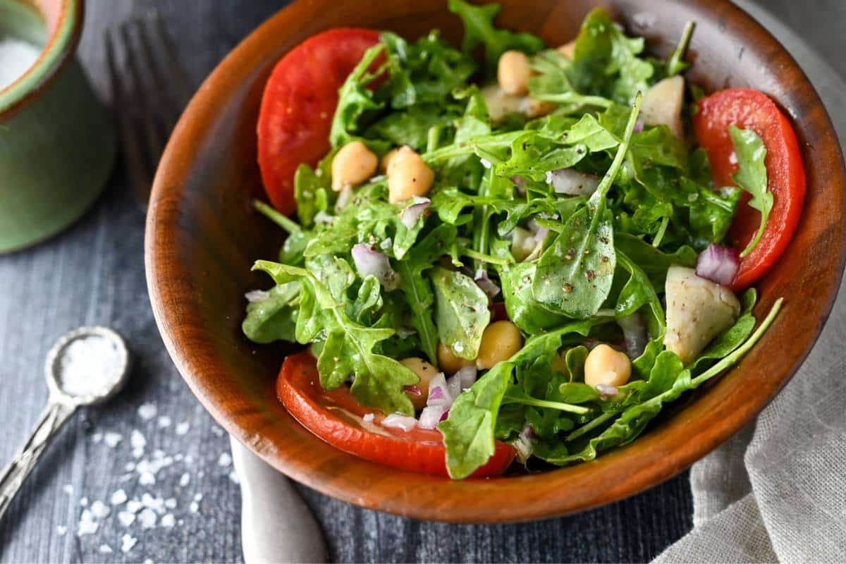 a close up photo of salad in a wood bowl with arugula, tomatoes, chickpeas, and artichokes