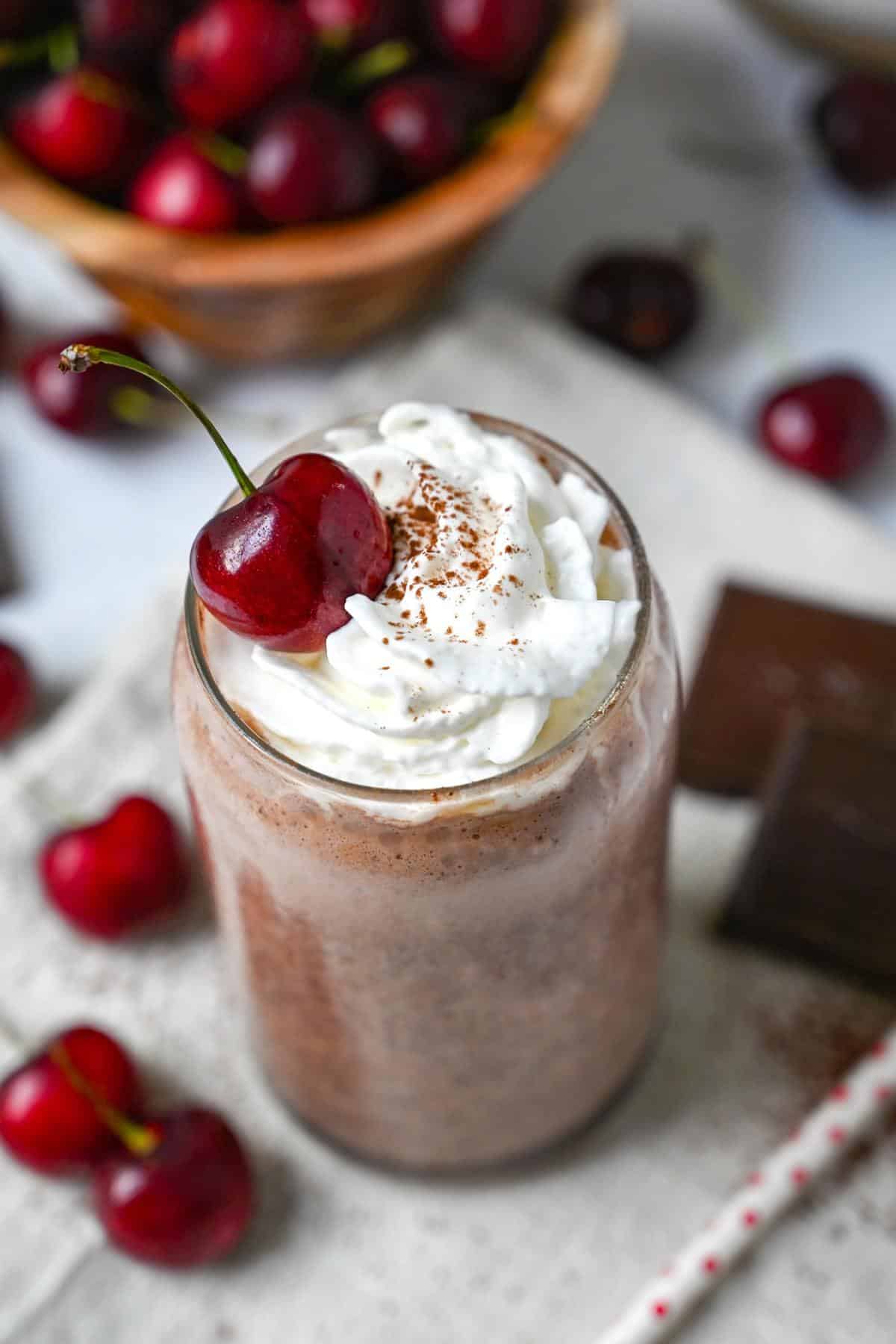 a cherry chocolate protein shake in a glass topped with whipped cream, a dusting of cocoa and a cherry