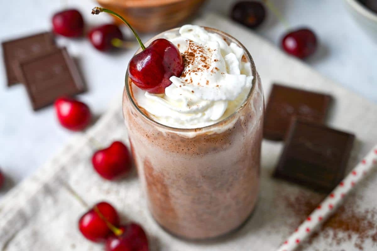 side view of a protein shake in a glass with whipped cream and cherry on top with cherries and dark chocolate all around it