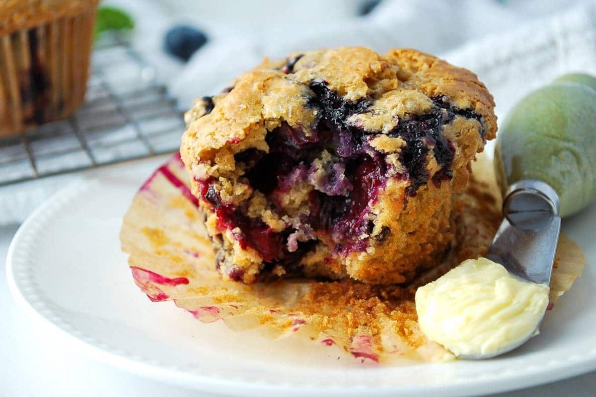 a blueberry muffin on a plate with a bite out of it with a knife with butter on it