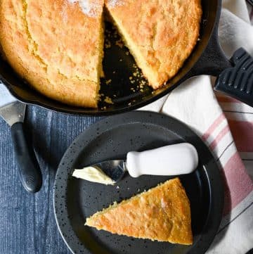 overhead photo of a cast iron skillet of cornbread with a slice cut out on a plate with a butter knife with butter on it