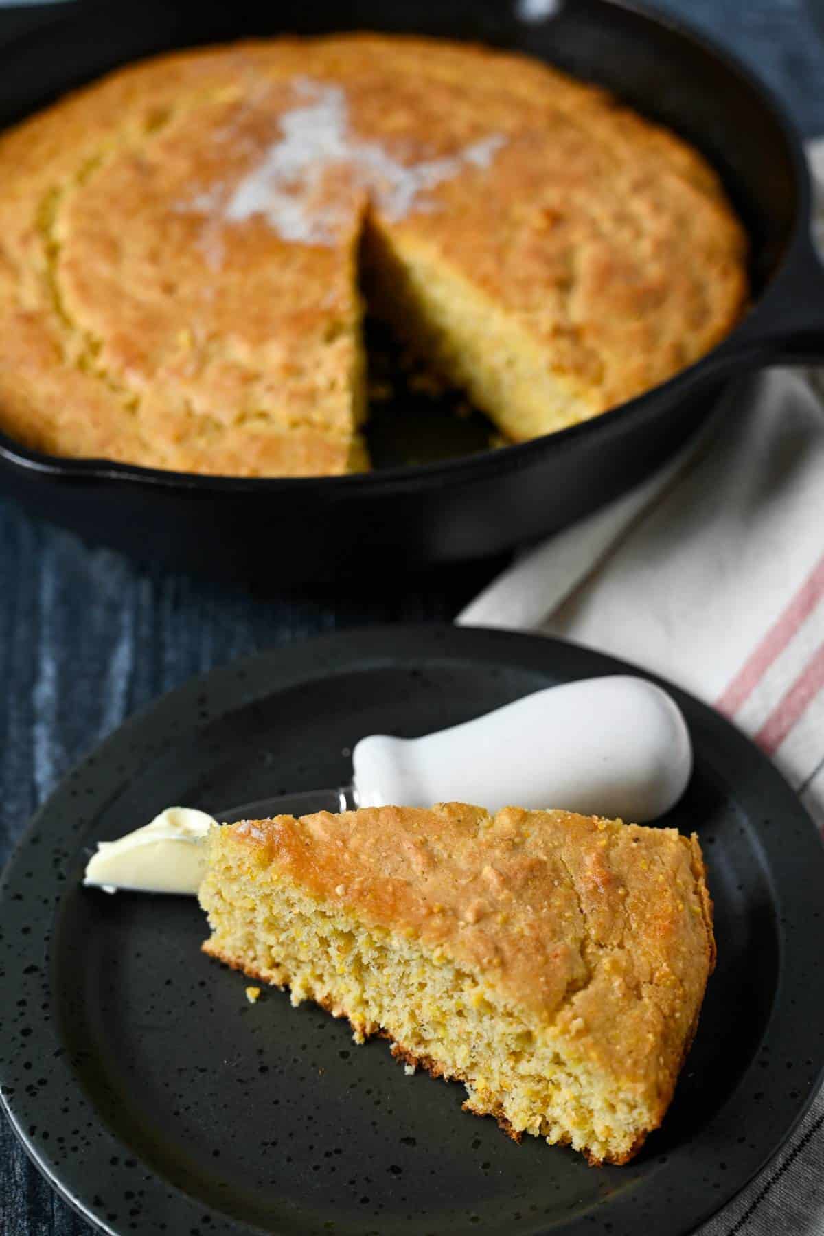slice of cornbread on a plate with a cast iron skillet of cornbread behind it