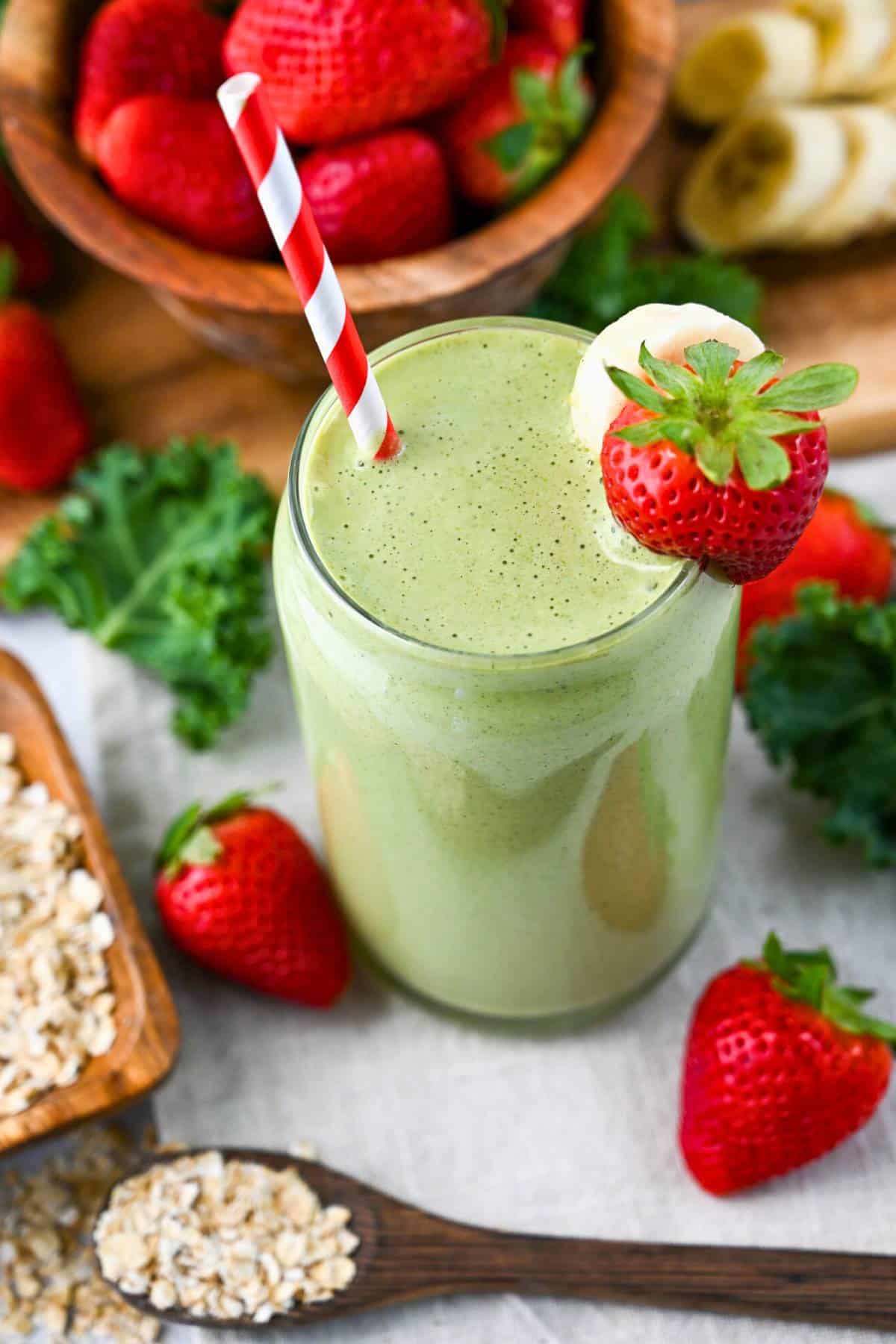 a green smoothie in a glass with a straw, a dish of oats with a spoon and fresh strawberries and kale