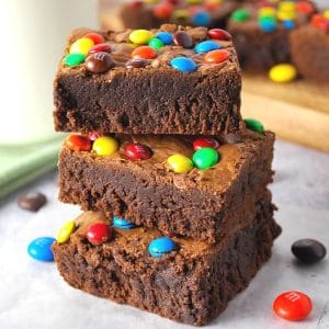 stack of fudgy protein brownies with m&ms with a tray of brownies and glass of milk in the background