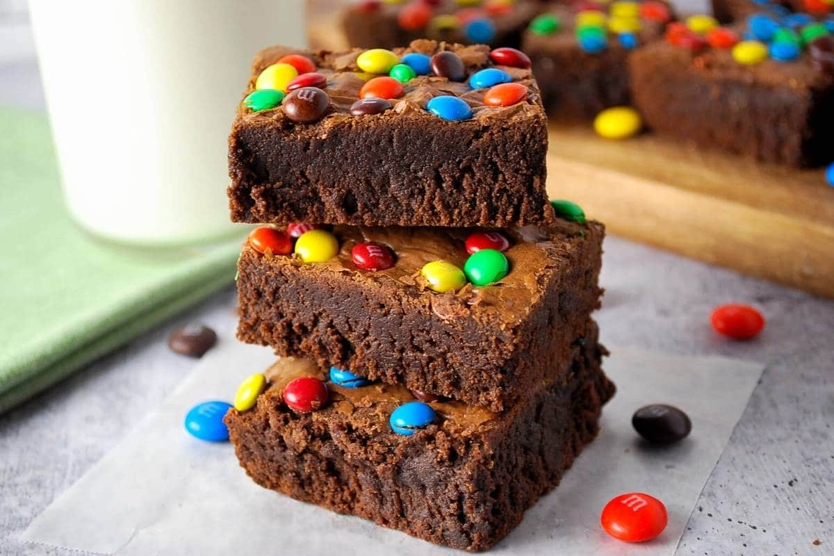 a stack of three brownies with m&m's on top with a glass of milk and tray of brownies