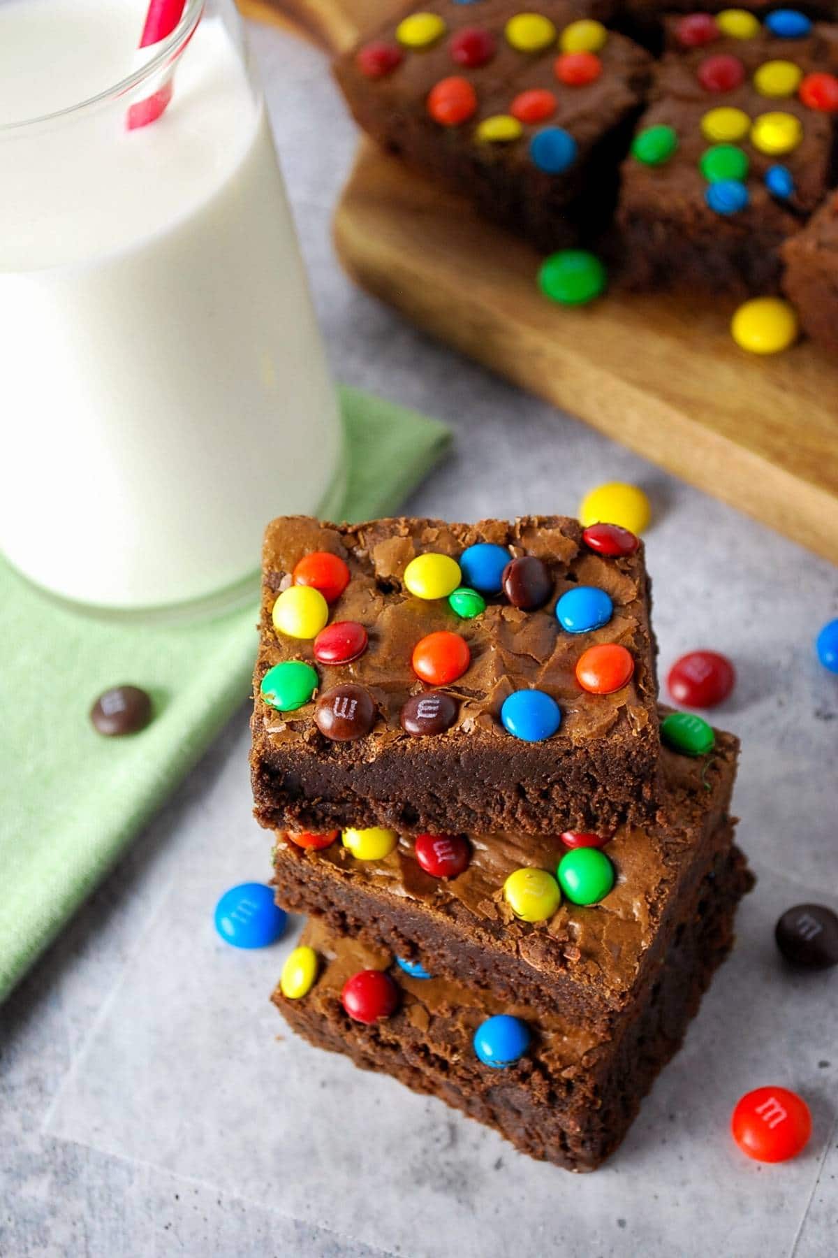 stack of fudgy protein brownies next to a glass of milk with a red striped straw with m&ms sprinkled around 