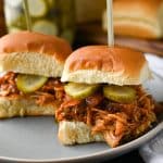 close up of two barbecue chicken sliders on a gray plate with a jar of pickles behind it