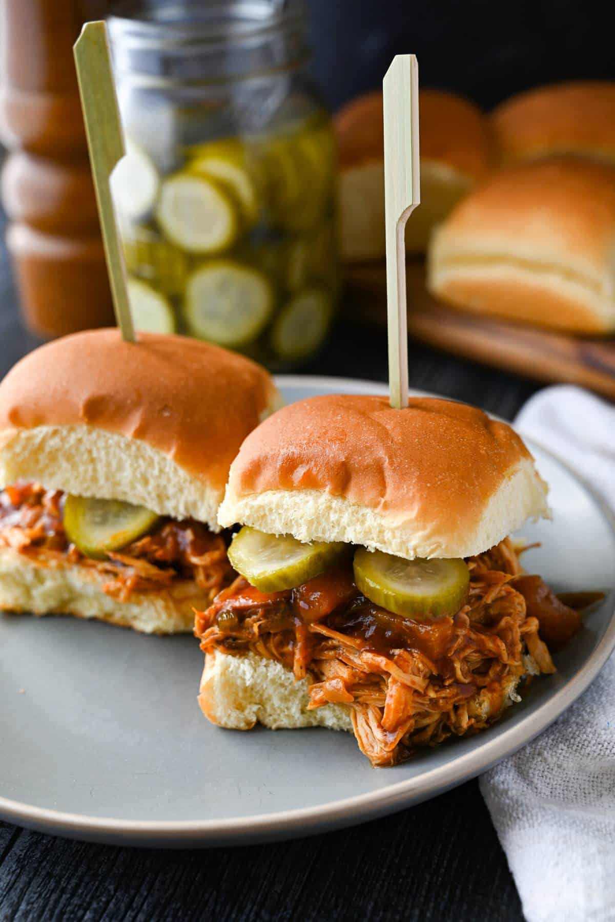 shredded barbecue chicken on two slider buns with pickles and a platter of buns behind it
