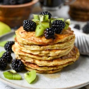 a zoomed in photo of a tall stack of banana cottage cheese pancakes dripping with syrup and topped with blackberries and kiwi with a fork on a plate