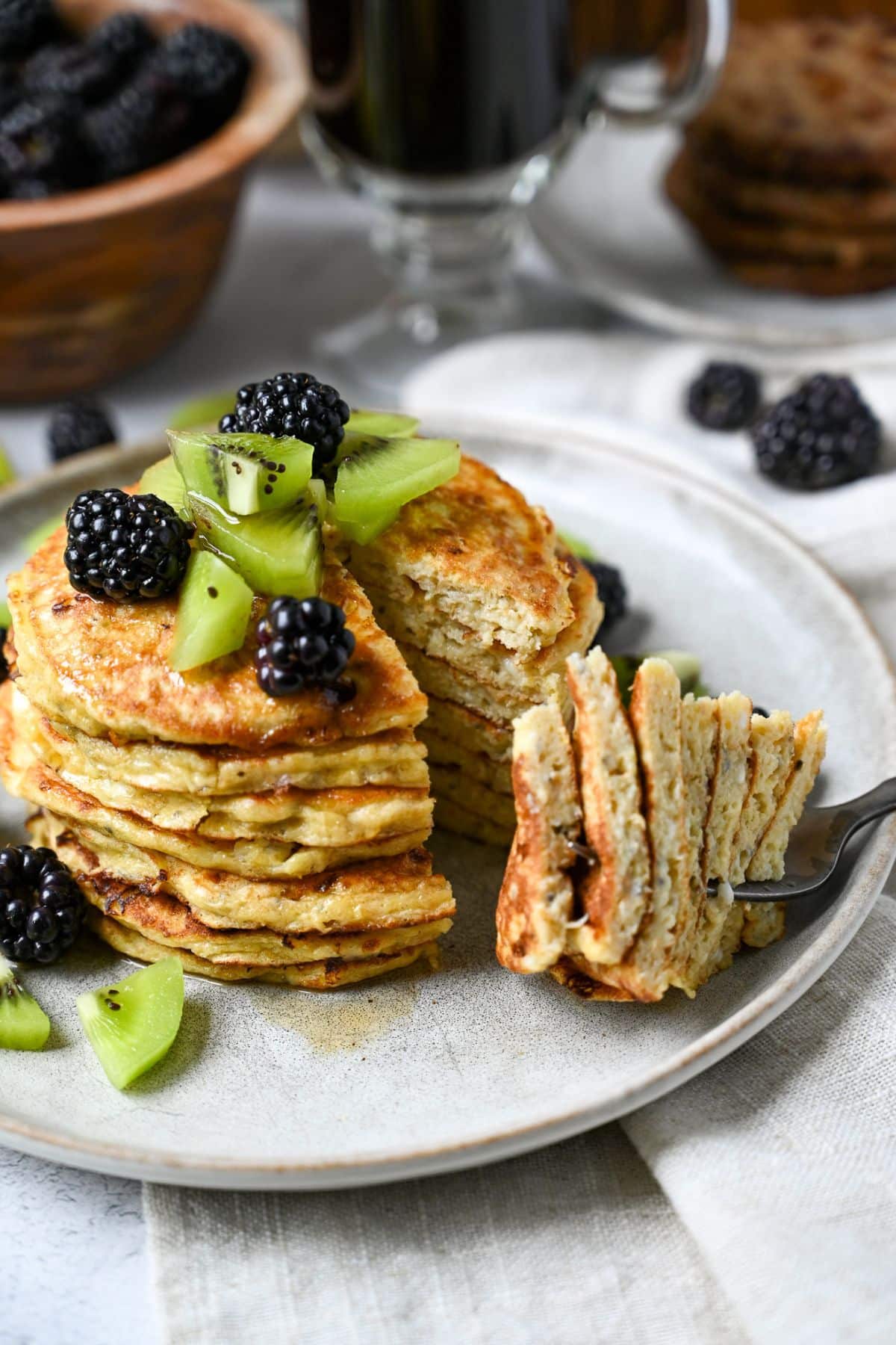 a stack of pancakes with a bite cut out on a fork with berries and kiwi