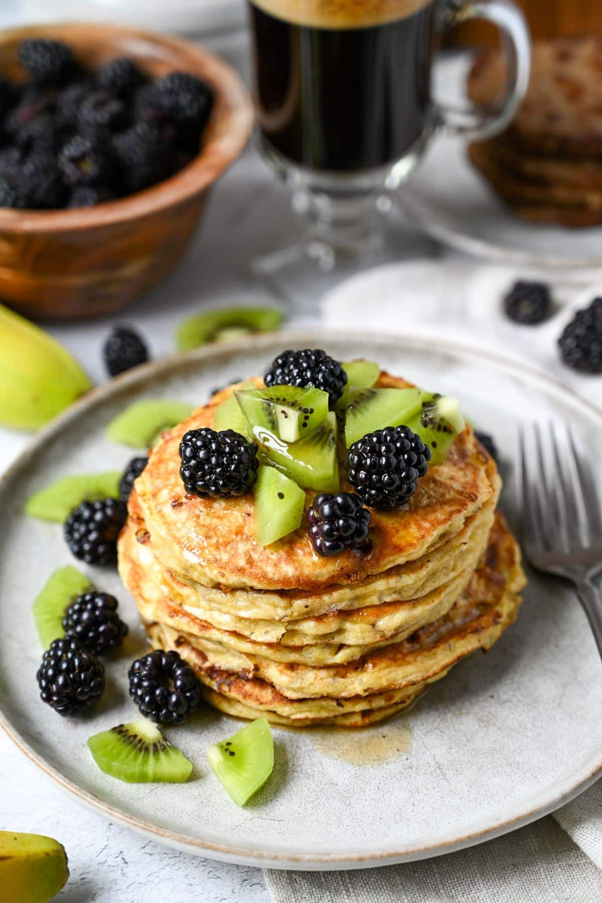 a plate of pancakes with kiwi and blackberries with a bowl of blackberries and a cup of espresso behind it