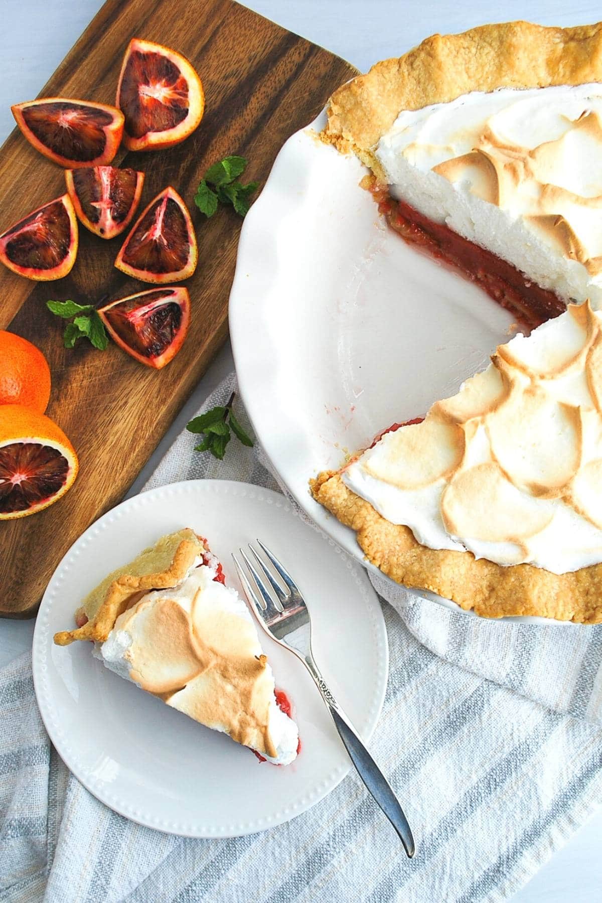 overhead photo of an orange meringue pie with a slice on a plate with sliced blood oranges on a cutting board