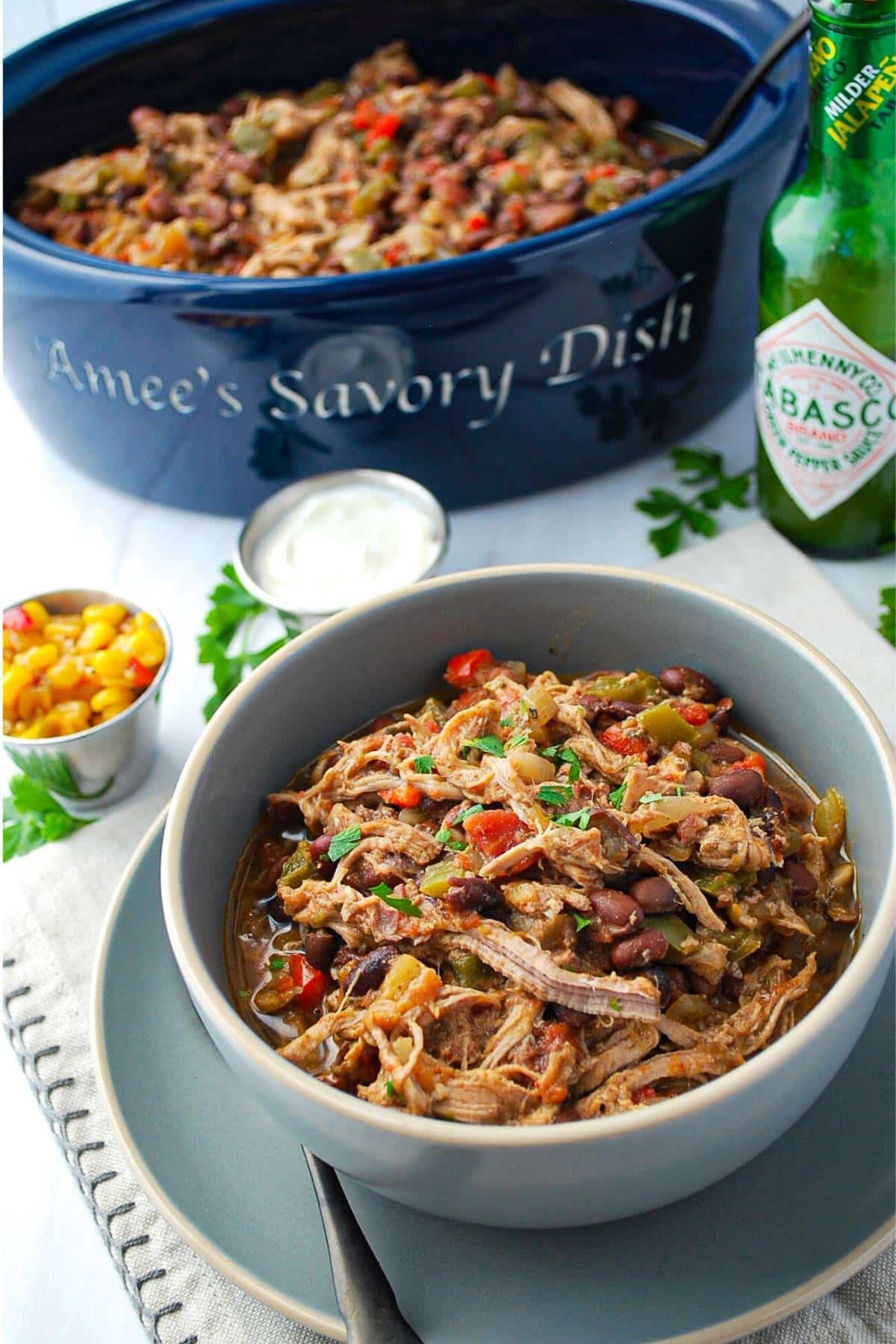 a serving bowl of chili with a bowl of chili in front of it with garnishes and a bottle of Tabasco