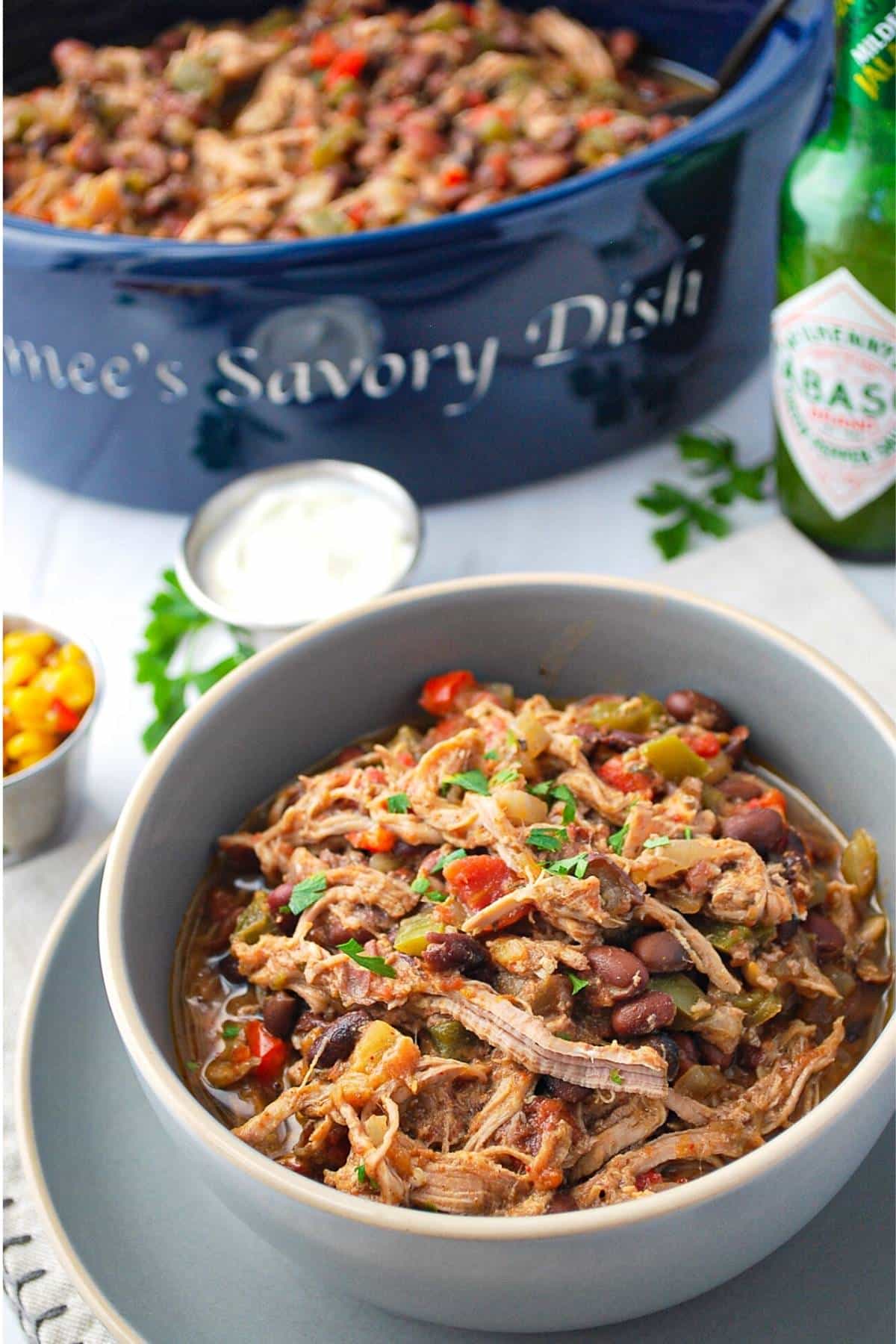 a bowl of pork tenderloin chili with a pot of chili in the background with sides of corn salsa and sour cream