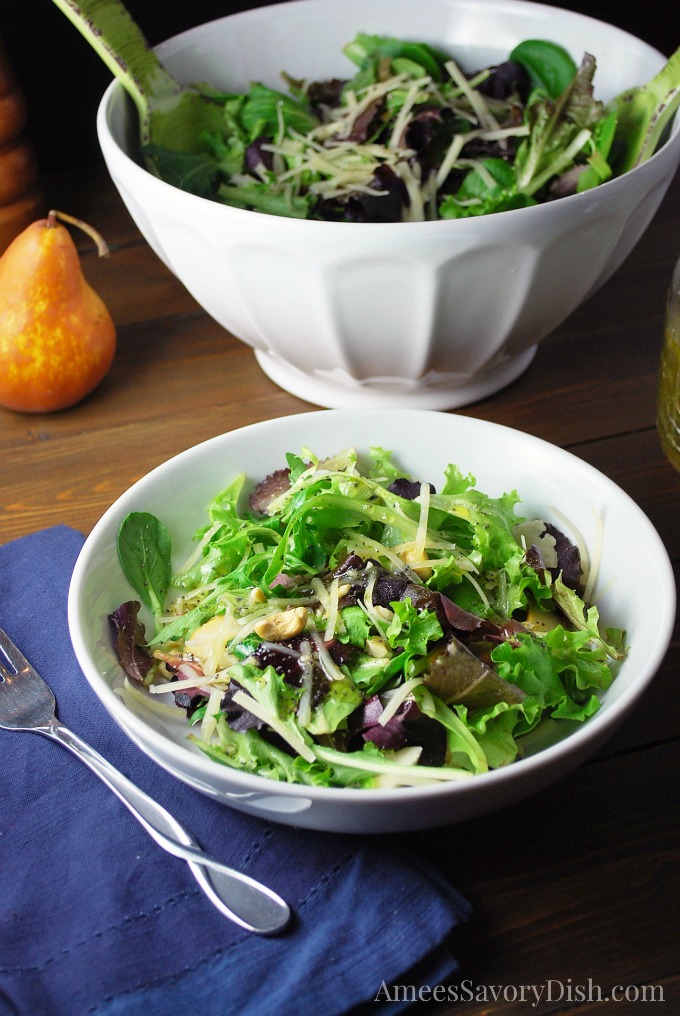 Mixed green salad with fruit in a white bowl with a blue napkin underneath and fork next to the bowl. A pear and bowl of salad is in the background