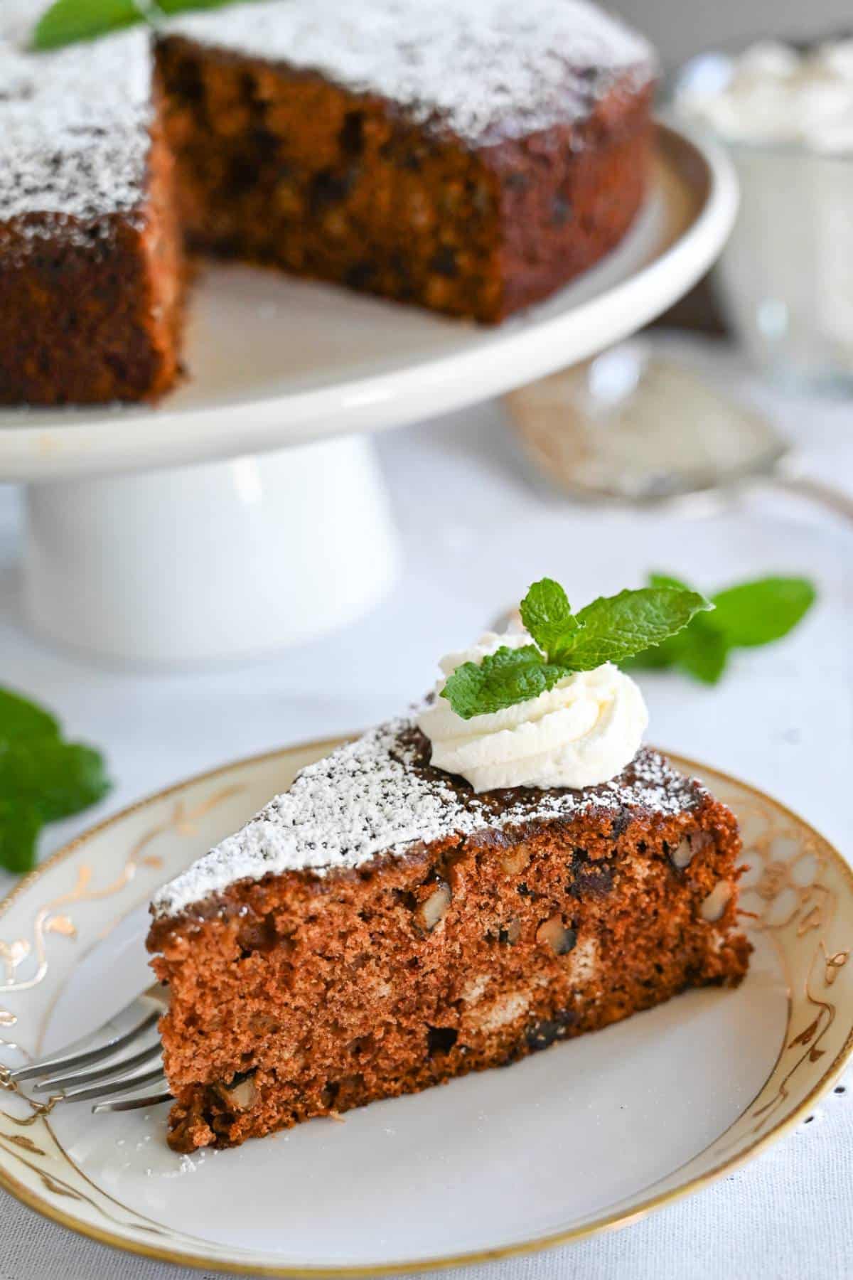 a slice of date walnut cake with whipped cream and a mint leaf with a cake on a platter behind it