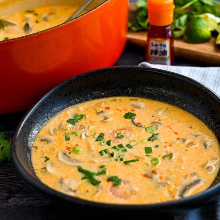 tom kha shrimp soup in a black bowl with a pot of soup and chili oil bottle behind it