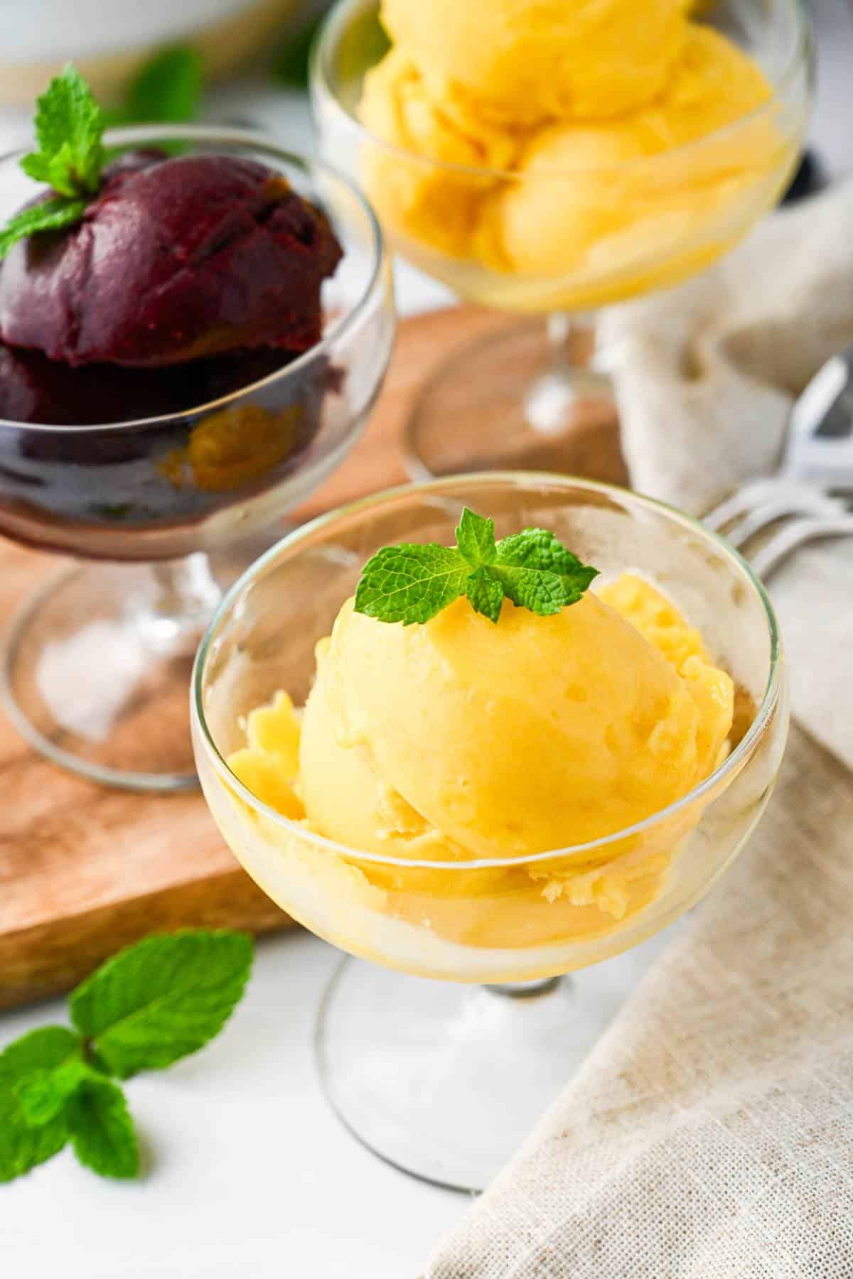 Three parfait dishes with a scoop of sorbet in them, one blueberry peach and two pineapple mango with fresh mint