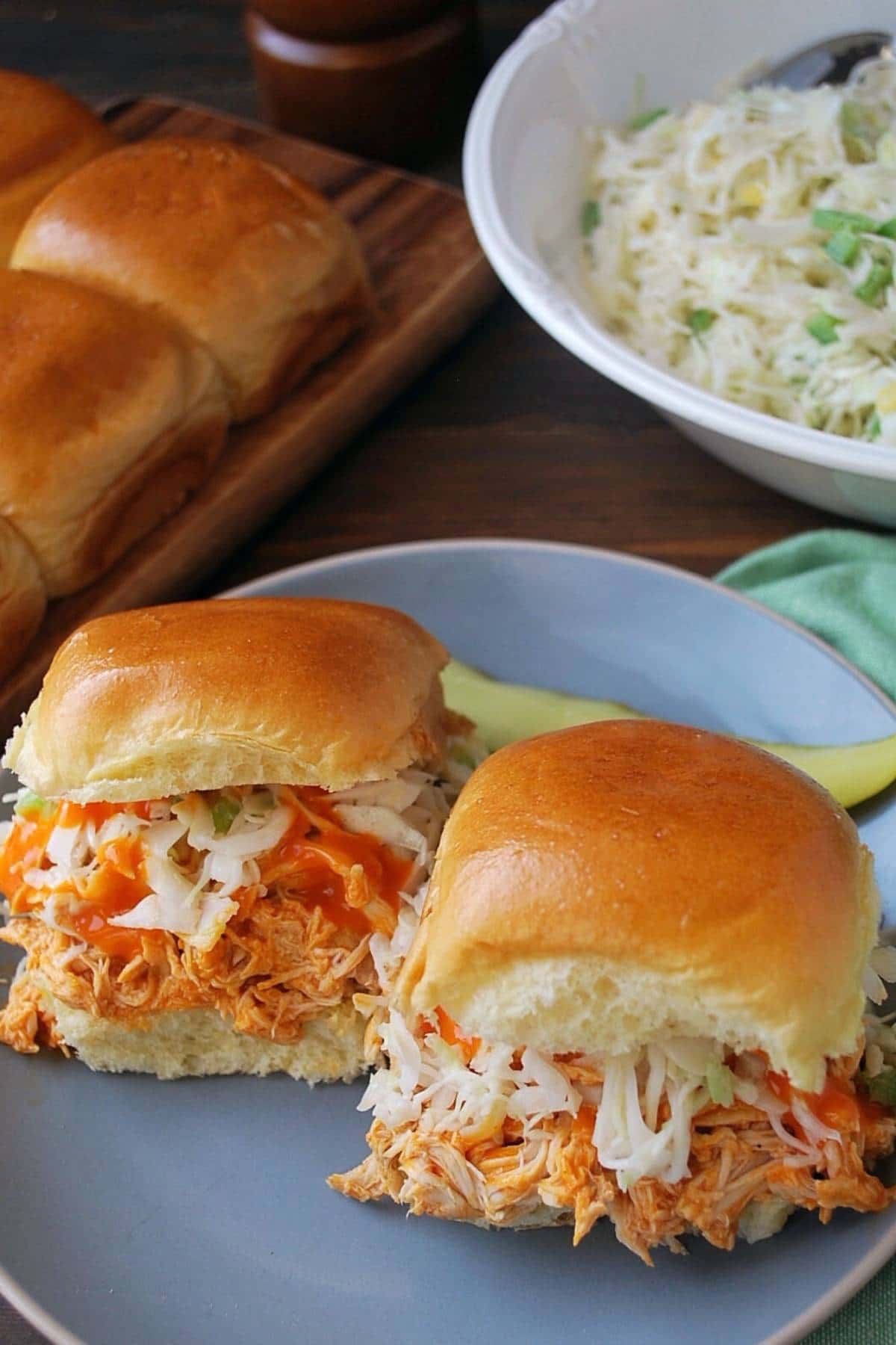 two buffalo chicken sliders topped with slaw and hot sauce with a bowl of slaw in the background
