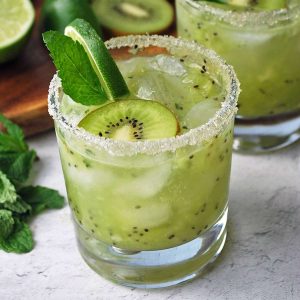 two kiwi mojitos in glasses with a sugared rim and garnished with lime, mint, and a slice of kiwi fruit