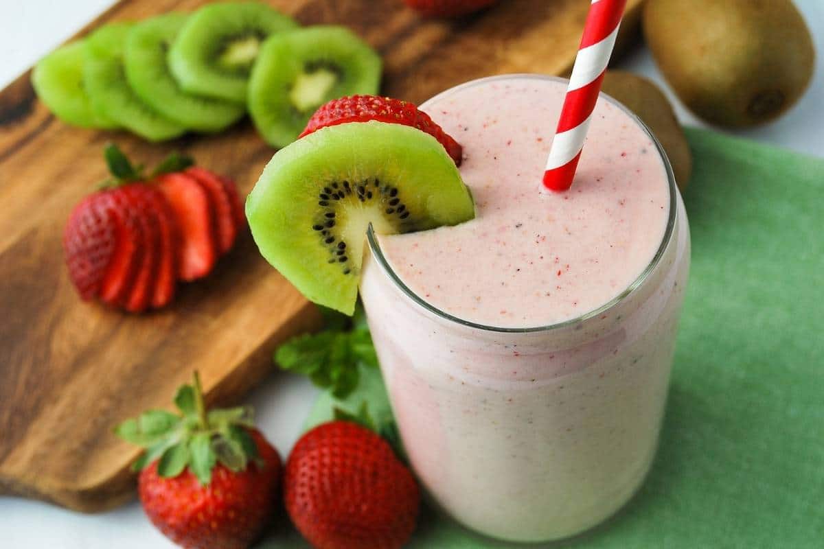strawberry kiwi banana smoothie in a glass with sliced fruit and a cutting board with kiwi and berries in the background