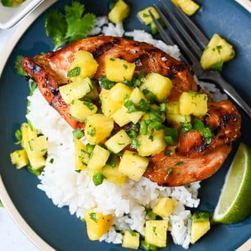 close up of grilled teriyaki chicken topped with jalapeno pineapple salsa over rice on a blue plate with a fork