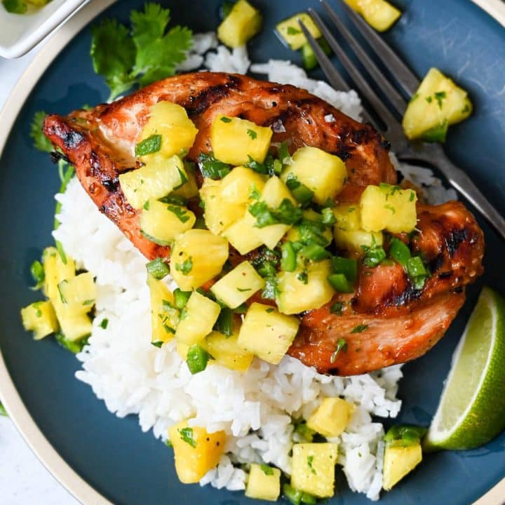 close up of grilled teriyaki chicken topped with jalapeno pineapple salsa over rice on a blue plate with a fork