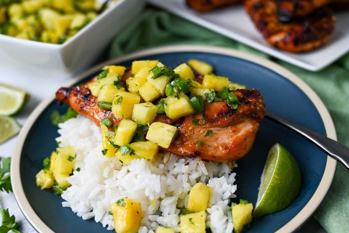 side view of grilled chicken topped with pineapple salsa and rice on a plate with a fork with a green napkin