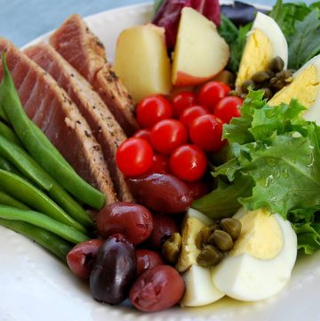 close up of sliced tuna, green beans, boiled eggs, olives, sliced potatoes, greens and capers in a bowl