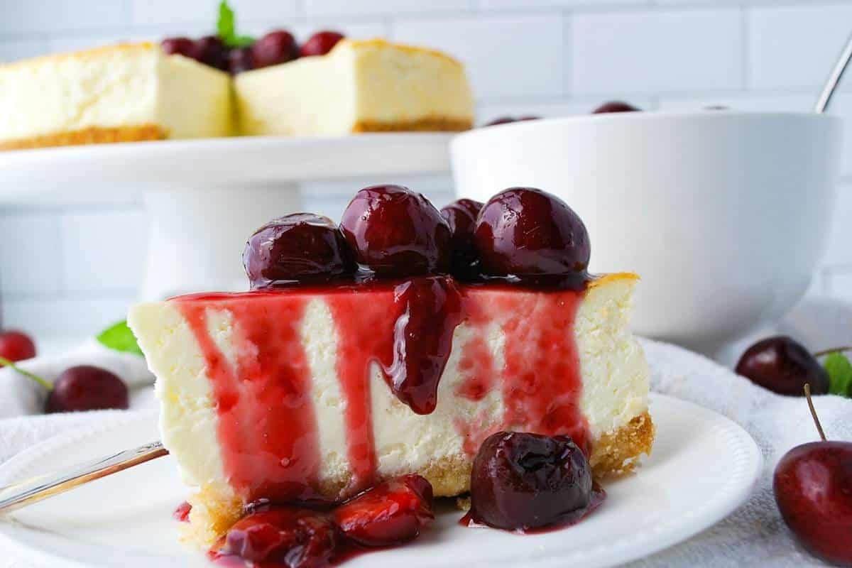 looking up at a slice of lemon cheesecake with cherry compote with a bowl of compote and platter of cheesecake behind it