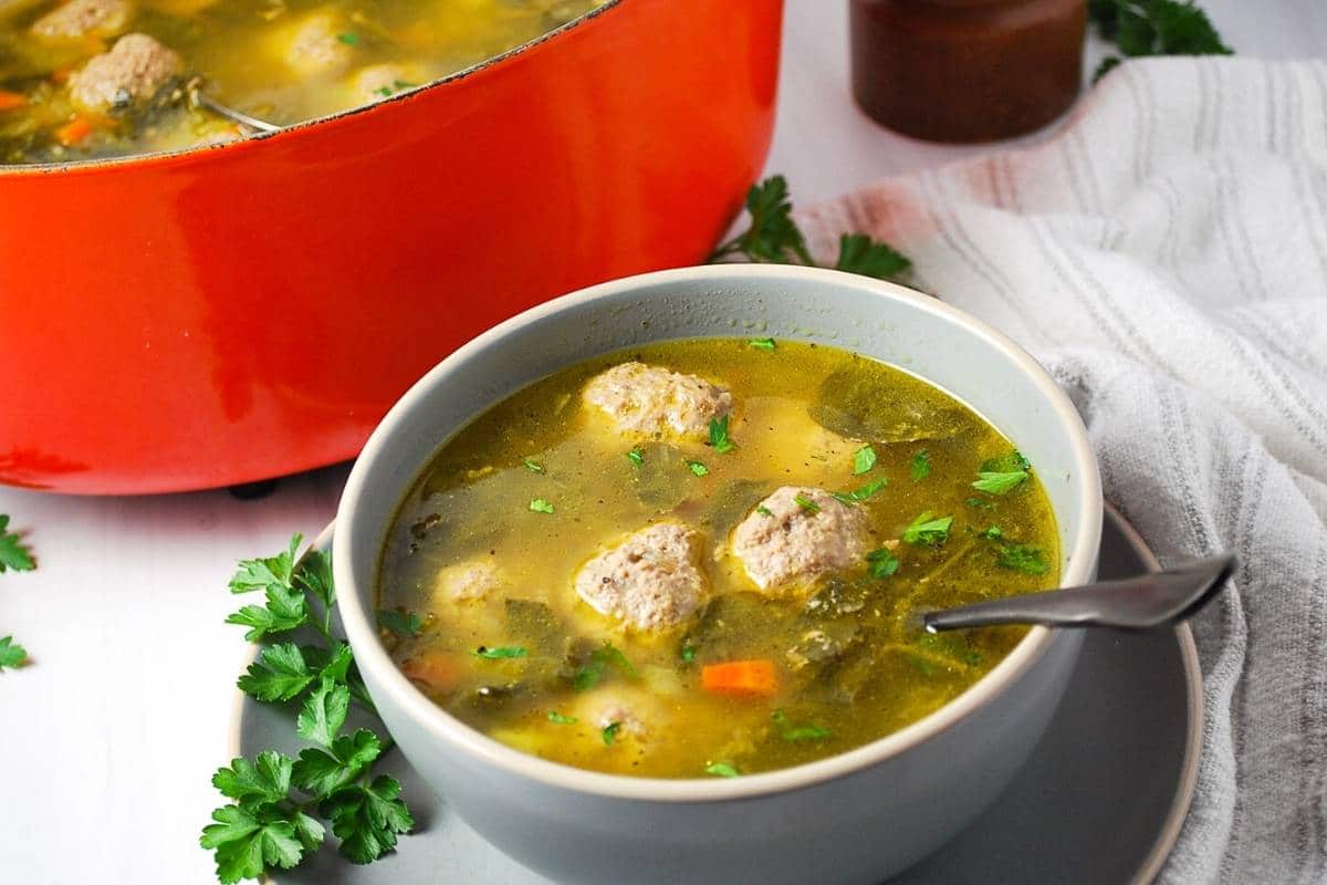 a bowl of meatball soup with herbs around it and a spoon