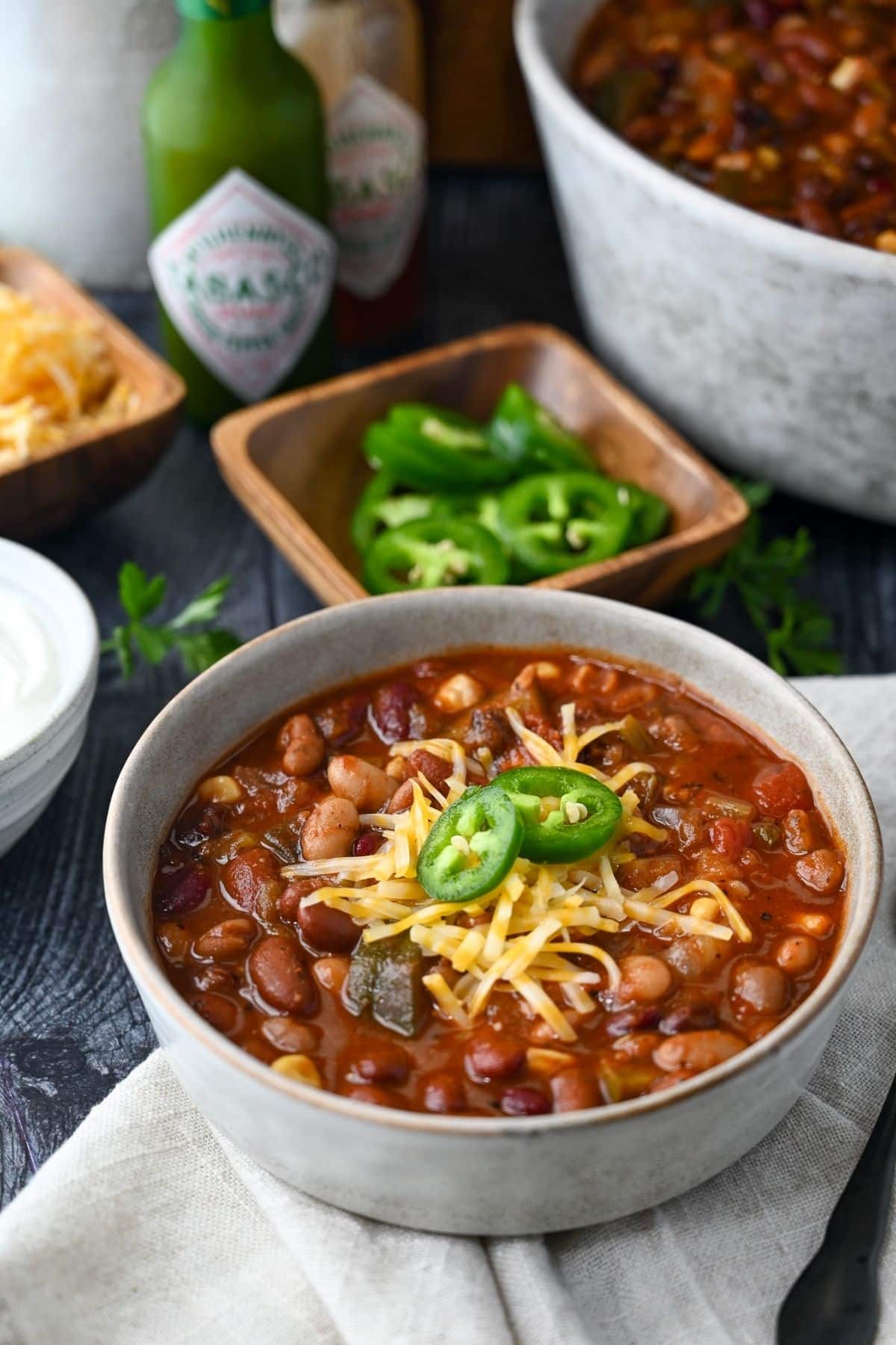 a bowl of veggie chili topped with cheese and slices of jalapenos with a bowl of jalapenos, hot sauce, and serving bowl of chili behind it