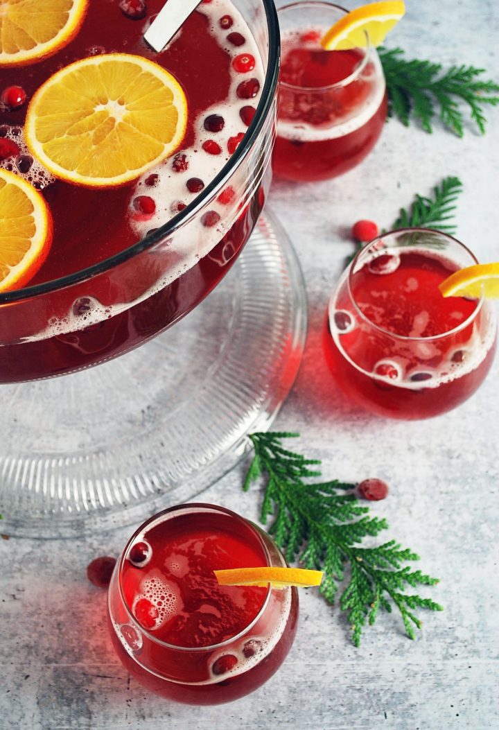 side angle photo of filled punch bowl garnished with oranges and cranberries with filled glasses on the side