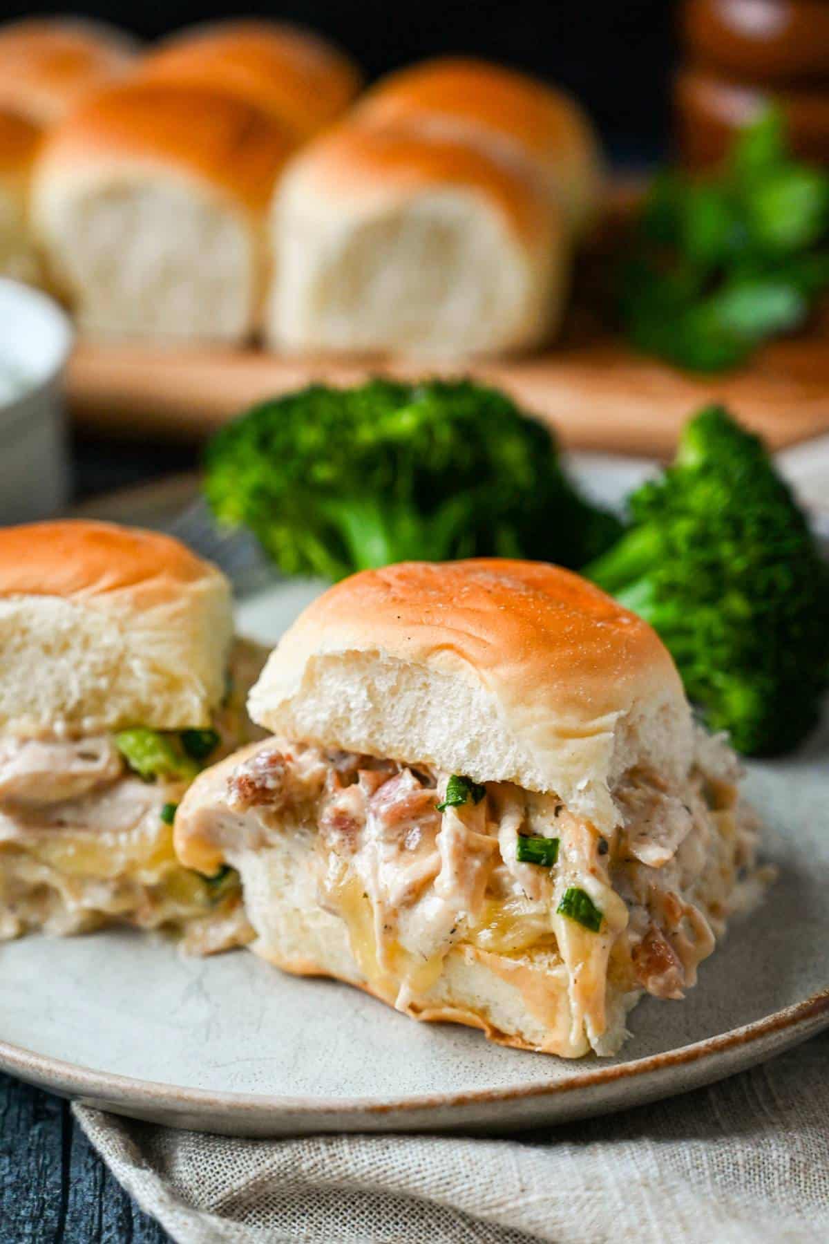 two crack chicken sliders on Hawaiian rolls with broccoli on the side and a platter of rolls in the background