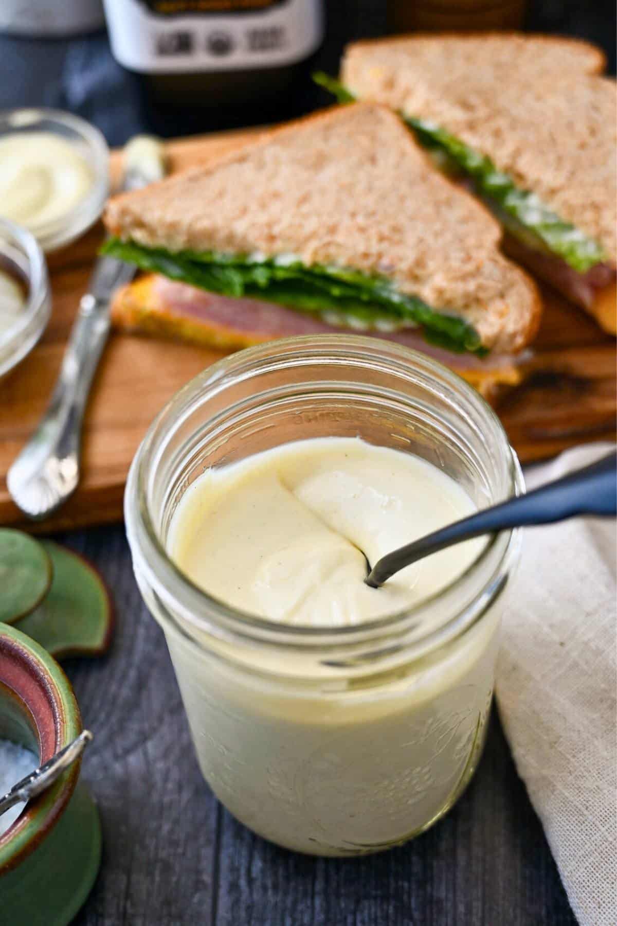 a mason jar of mayo with a spoon with a dish of mayo next to a sandwich on a cutting board