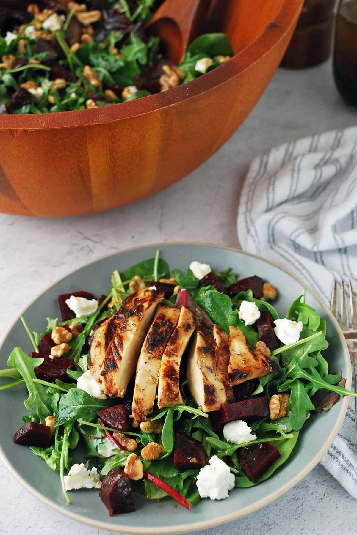 A mixed green salad topped with beets, goat cheese, walnuts, and grilled chicken on a plate with a salad bowl and napkin in the background