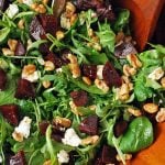 close up of a mixed green salad with beets, goat cheese, walnuts, and dressing being tossed together with a wooden spoon