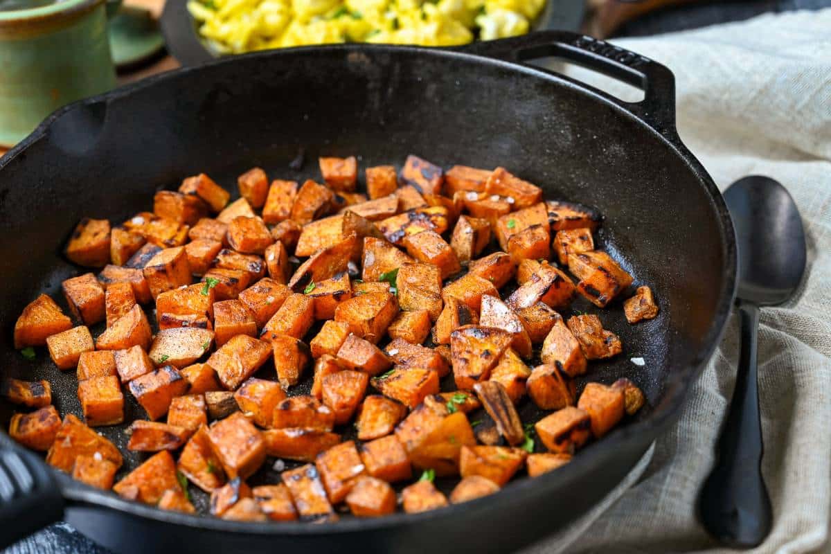 side view of a cast iron skillet with seasoned cooked sweet potatoes with a napkin beside it and a bowl of eggs behind it